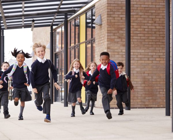Primary school kids, wearing school uniforms and backpacks, running on a walkway outside their school building, front view
