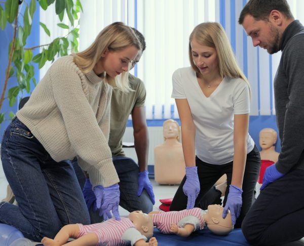 Group of people during the first aid training with instructor showing on manikin how to do artificial respiration for the baby.